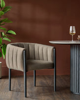 A contemporary chair with channel tufting and dark metal legs sits next to a round table with a fluted base.