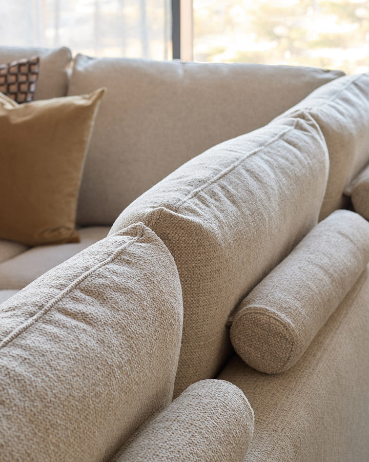 Beige fabric corner chaise sofa with rolled arms, close-up detail showing textured upholstery and piping seams.