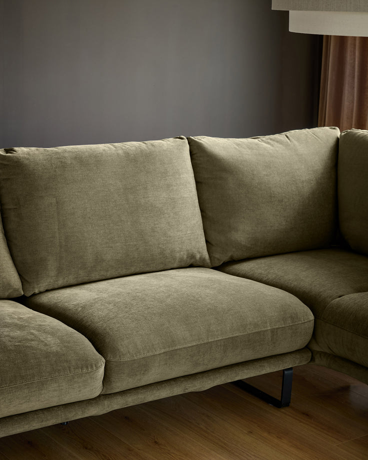 Olive green corner chaise sofa with right-hand chaise, dark legs, styled with patterned pillows in a neutral living room.