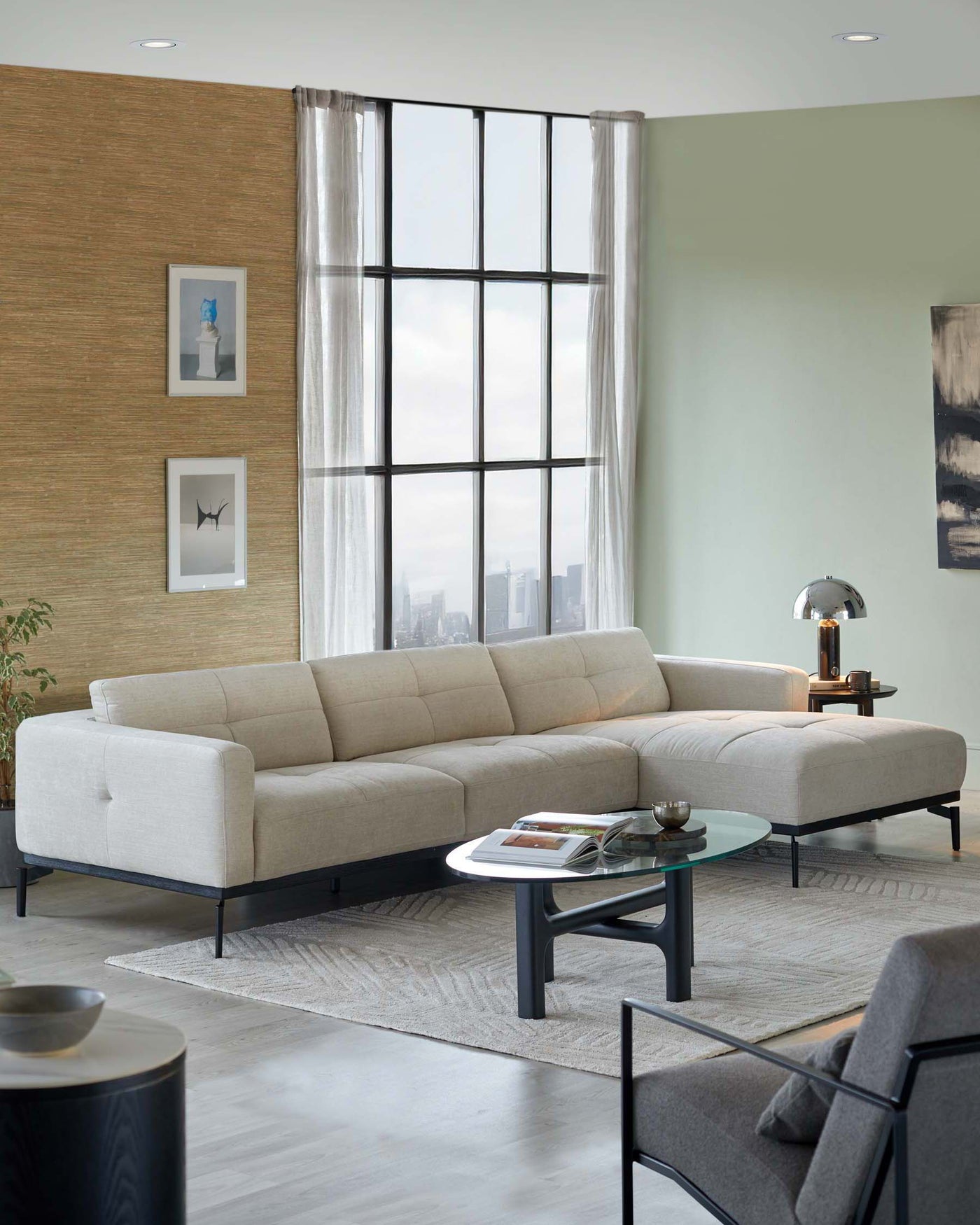 Modern beige L-shaped sectional sofa with clean lines and black metal legs, paired with an oval glass-top coffee table with a unique black base. A sleek, black floor lamp adds a touch of elegance to the space.