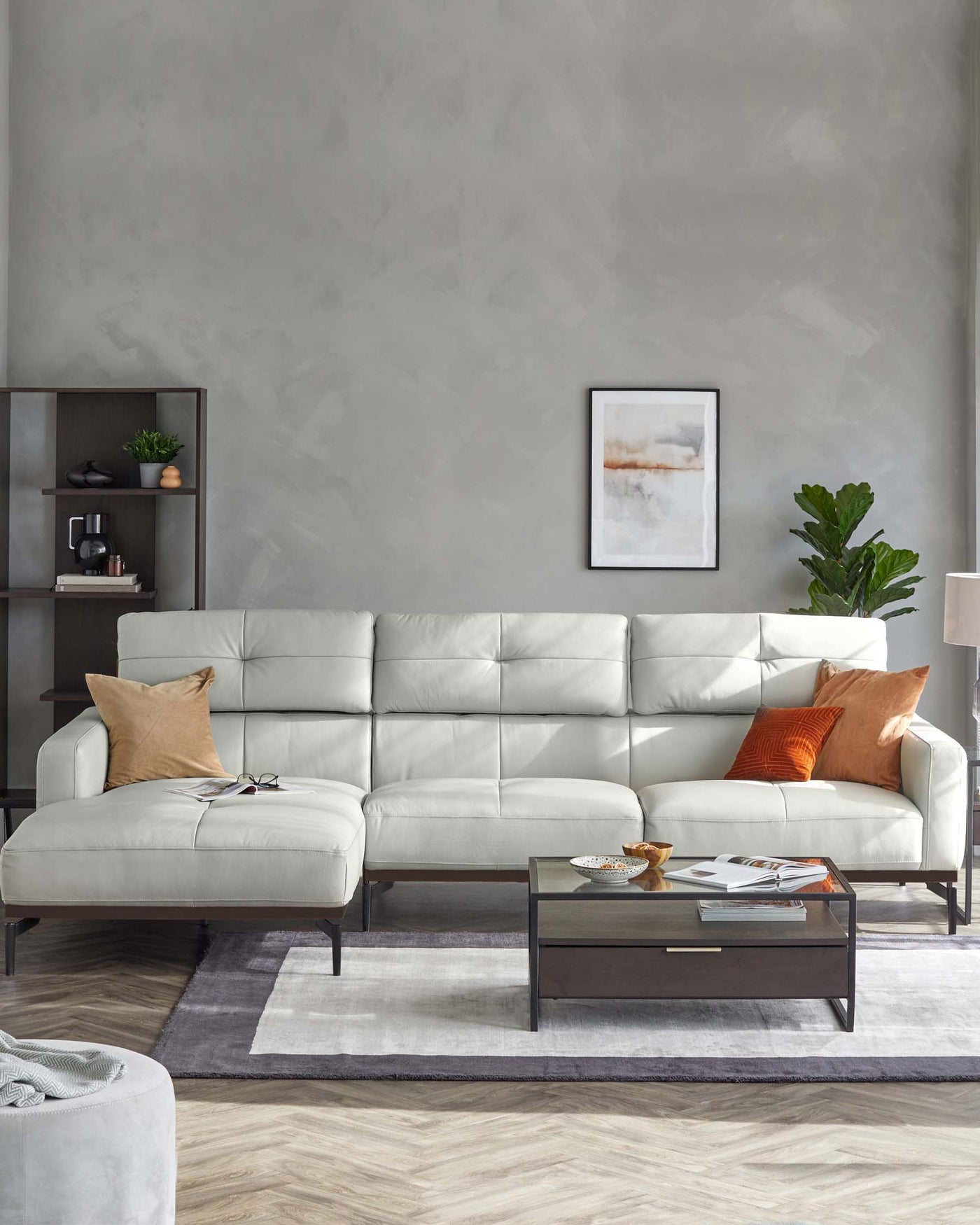 Modern living room setup featuring a sleek off-white L-shaped sectional sofa with tufted back cushions, accented with contrasting orange and beige throw pillows. In front of the sofa is a rectangular dark wood coffee table with a glossy finish and an open shelf, displaying decorative items and books. To the left, a minimalist dark wood shelving unit showcases sparse decorative pieces, enhancing the clean, contemporary aesthetic of the space.