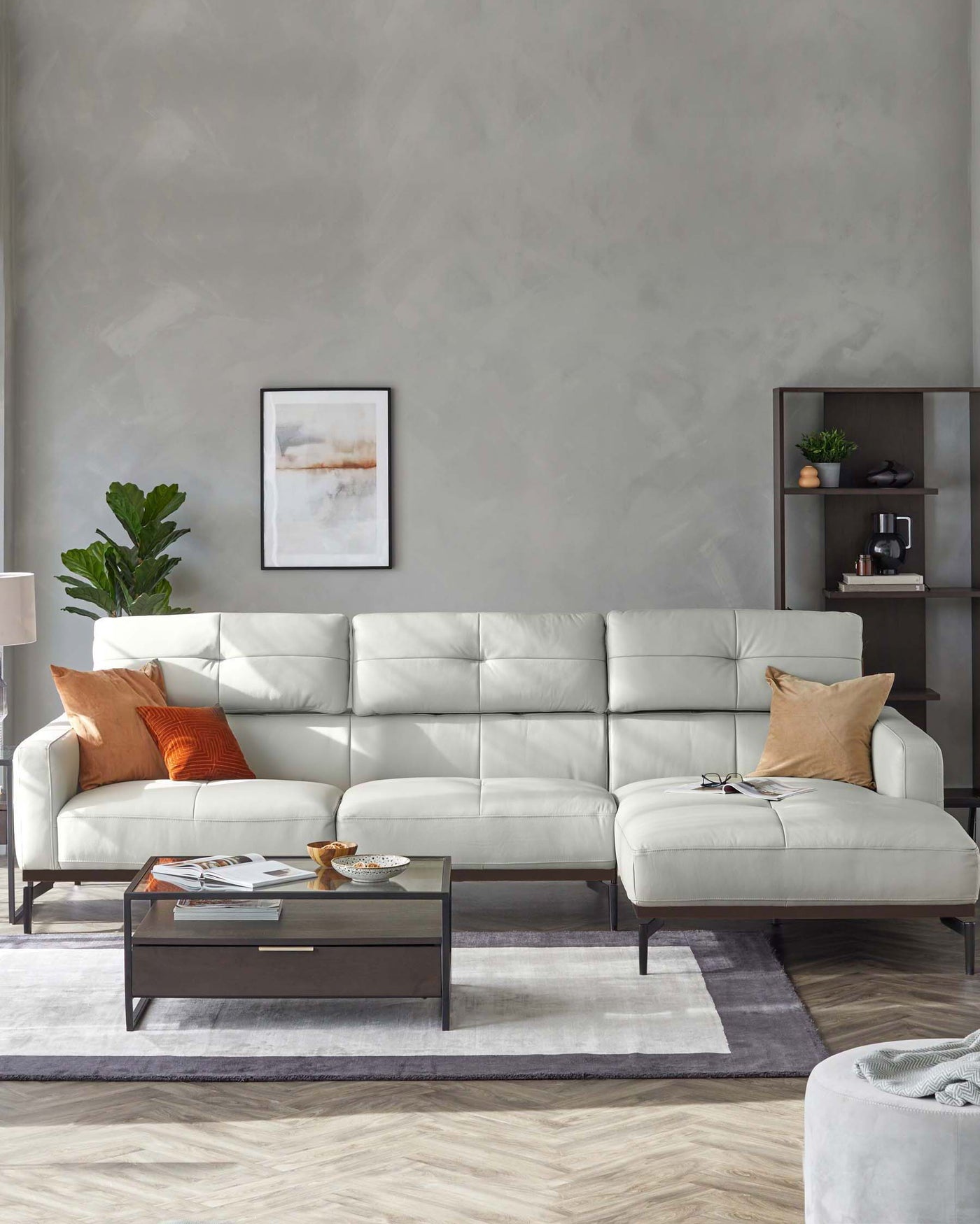 Modern light grey sectional sofa with a chaise lounge, accentuated with orange and tan throw pillows, a rectangular wooden coffee table with a glass top and a single drawer, set on a grey and white area rug. To the right, a dark wooden shelving unit displays decorative items and books, complemented by a white standing lamp and a green potted plant to the left.