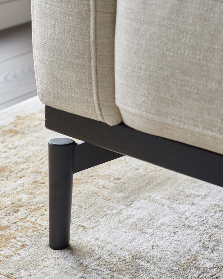 Close up of a neutral coloured sofa, with dark wooden legs, sat on a cream patterned rug.