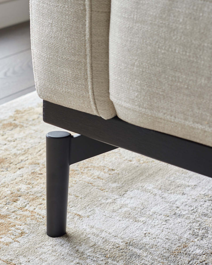 Close up of a neutral coloured sofa, with dark wooden legs, sat on a cream patterned rug.