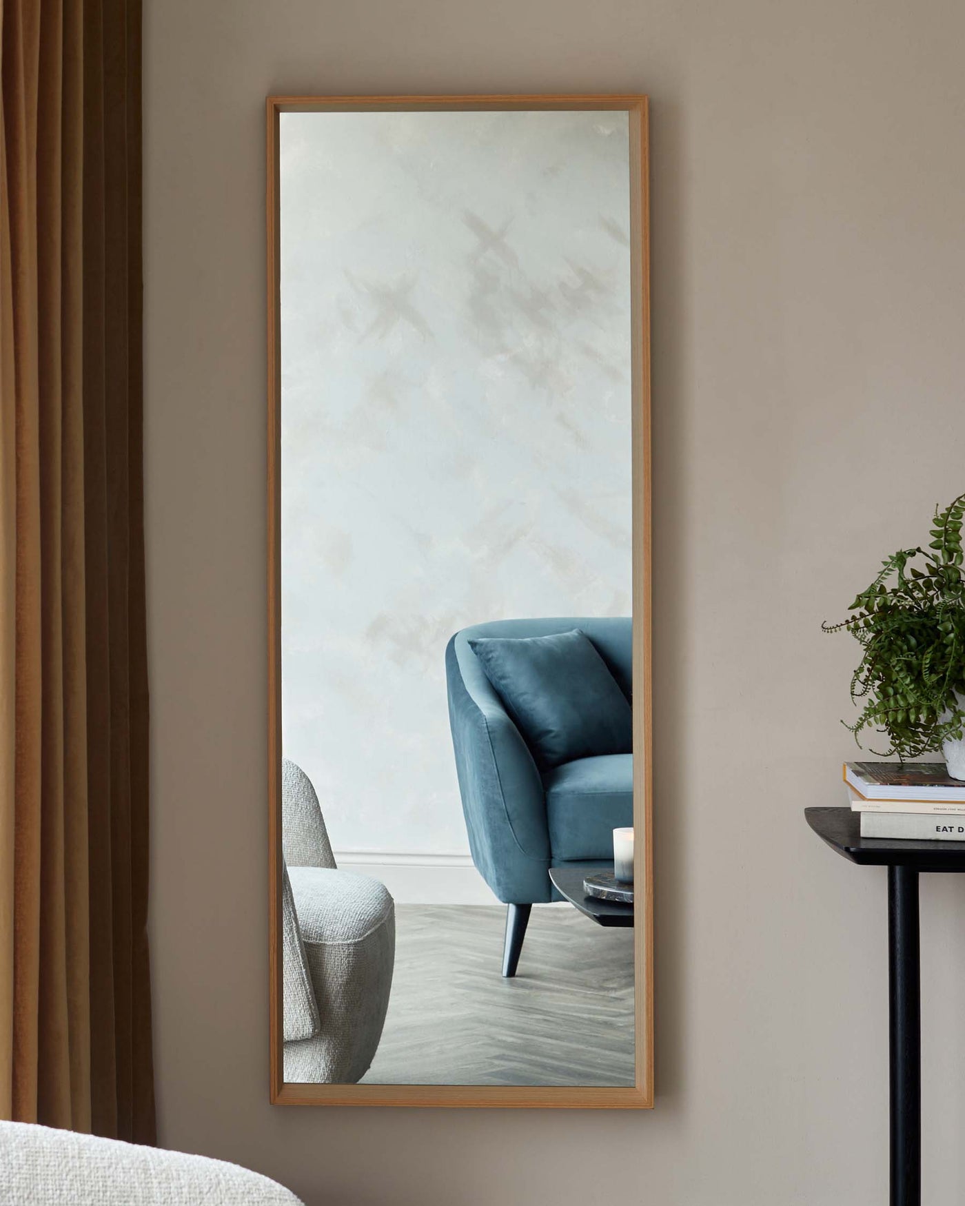 A modern teal blue armchair with a smooth, plush finish and angular black legs reflected in a large vertical mirror with a warm wooden frame. Beside it is a small black side table holding books and a potted plant, adding a cosy touch to the contemporary setting.