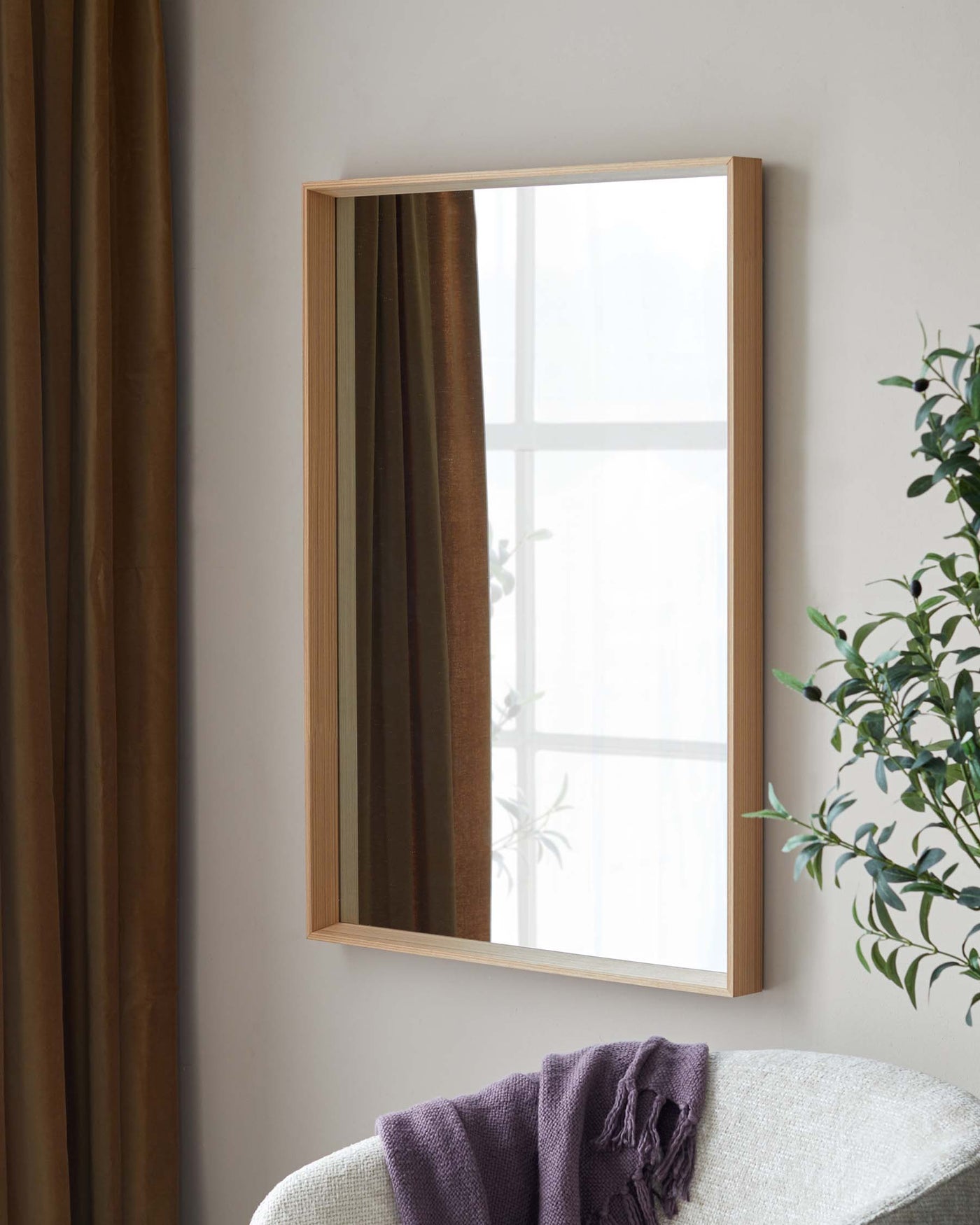 A modern, minimalist wooden-framed mirror mounted on a wall above an off-white, textured fabric armchair with a purple throw blanket casually draped over one armrest.