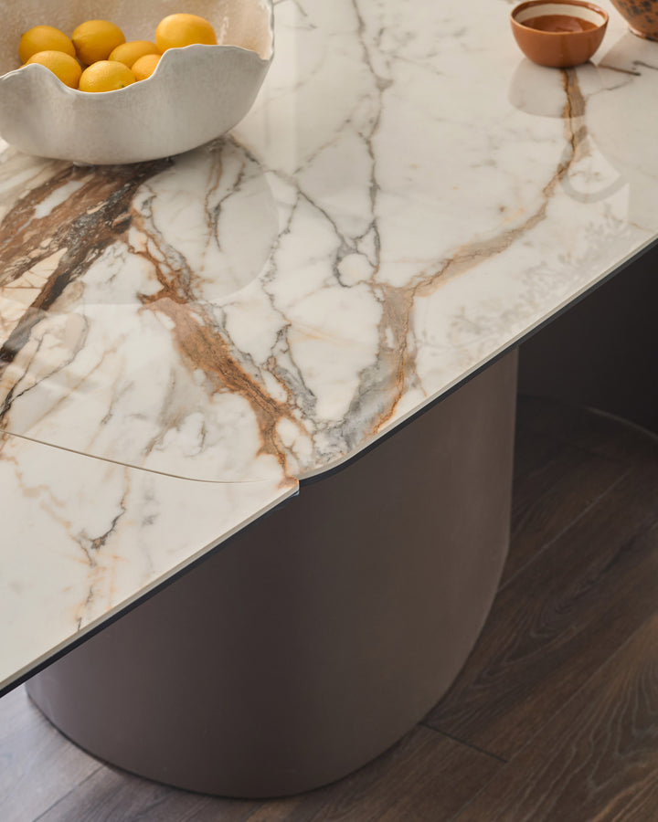 Cortona dining table with marble-effect ceramic top featuring brown and grey veining, curved dark base, styled with lemons and coffee.