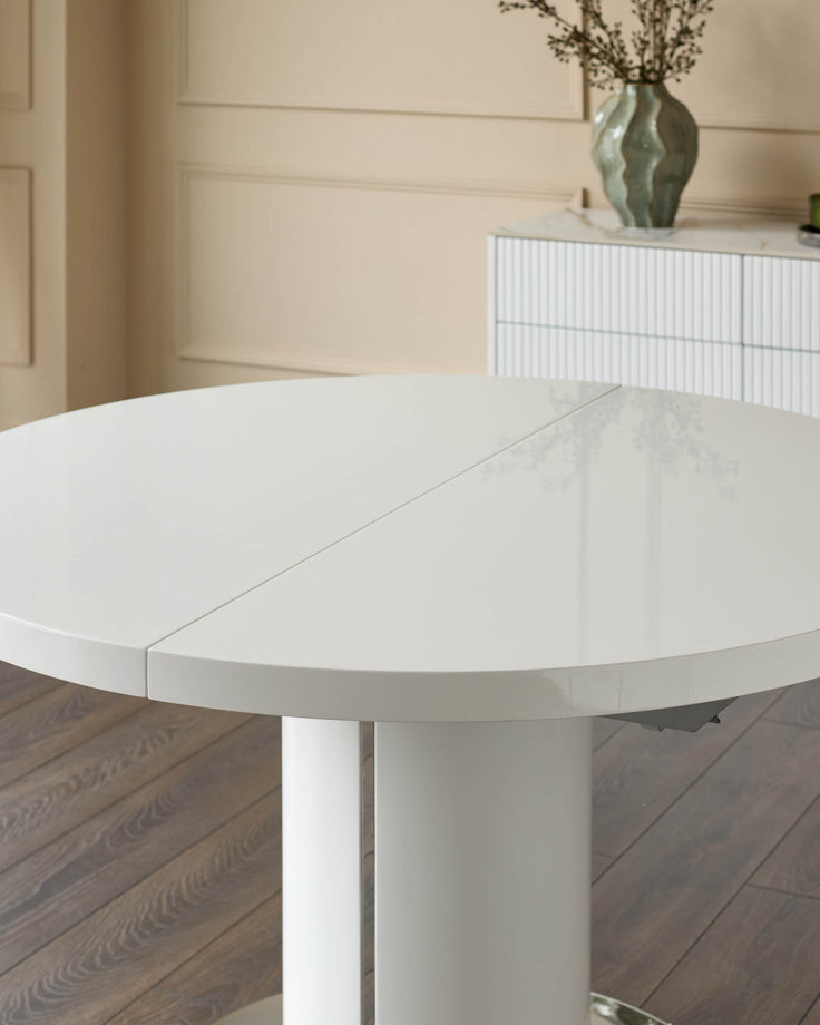 White gloss round extending dining table with cylindrical pedestal base, shown partially extended on wooden flooring.
