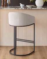 Dakota bar stool in natural fabric with black metal frame, curved back, footrest, shown against beige paneled wall.