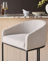 Dakota bar stool in natural fabric with curved back, black metal legs, shown in styled setting with marble countertop.