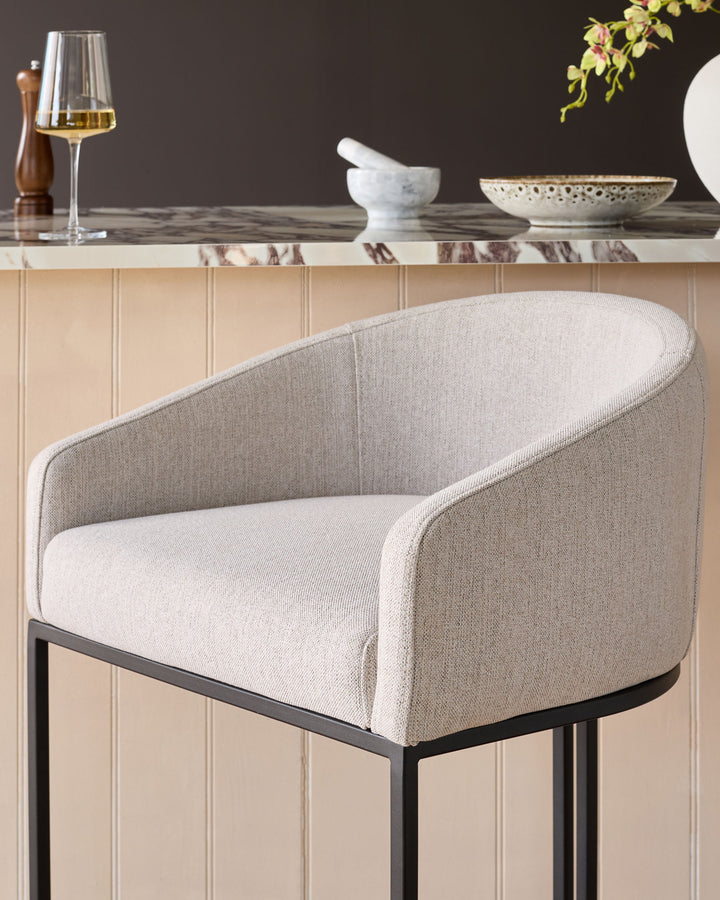 Dakota bar stool in natural fabric with curved back, black metal legs, shown in styled setting with marble countertop.