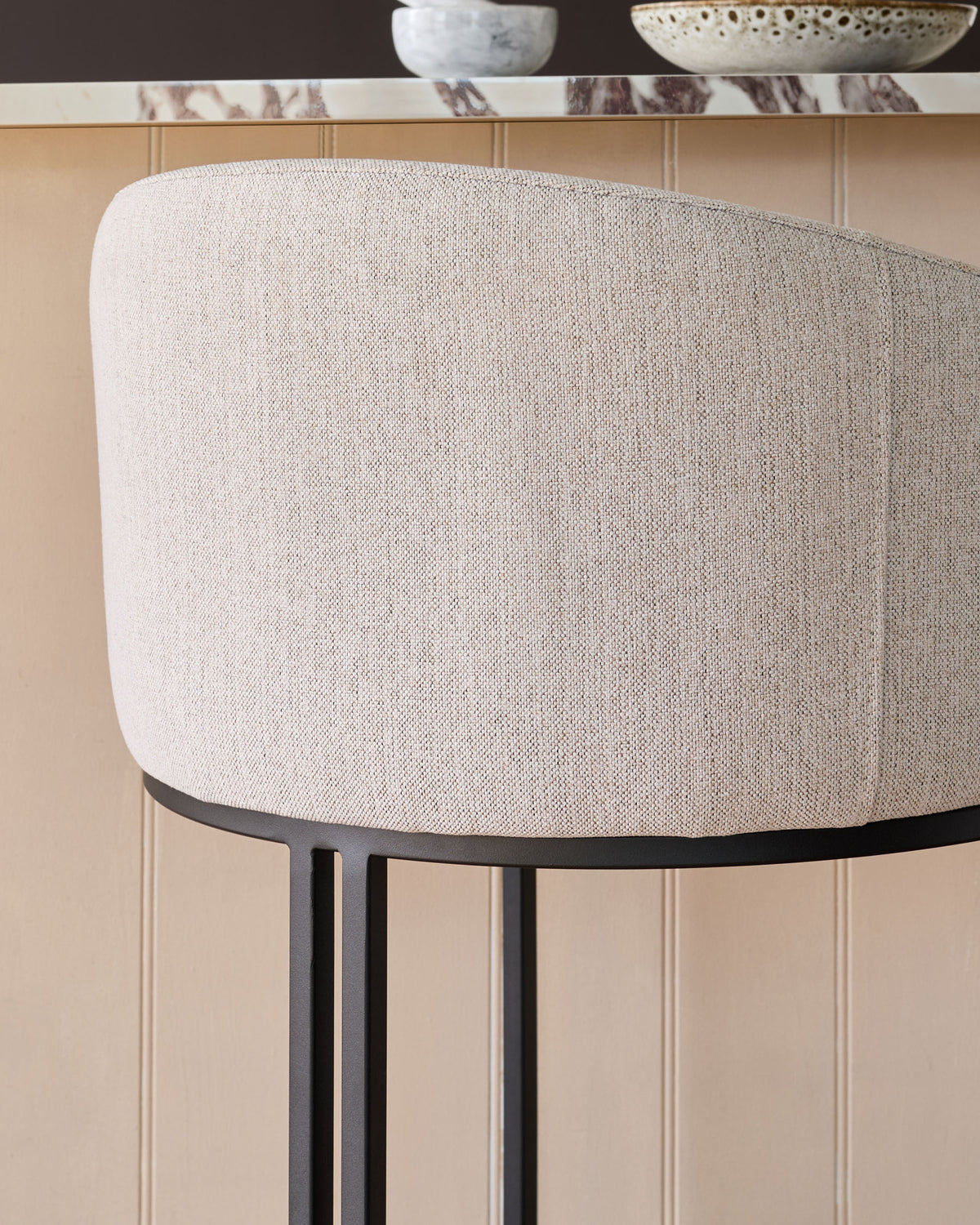 Dakota bar stool in natural fabric with black metal frame, shown from side angle against beige paneled wall.