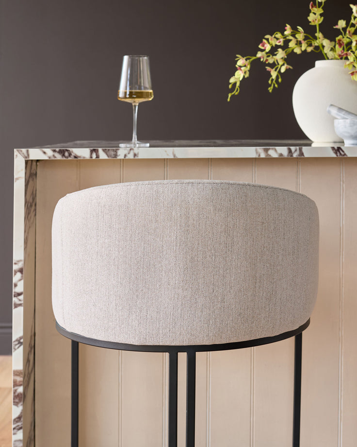 Dakota bar stool in natural fabric with black metal frame, shown against marble countertop in styled interior setting.