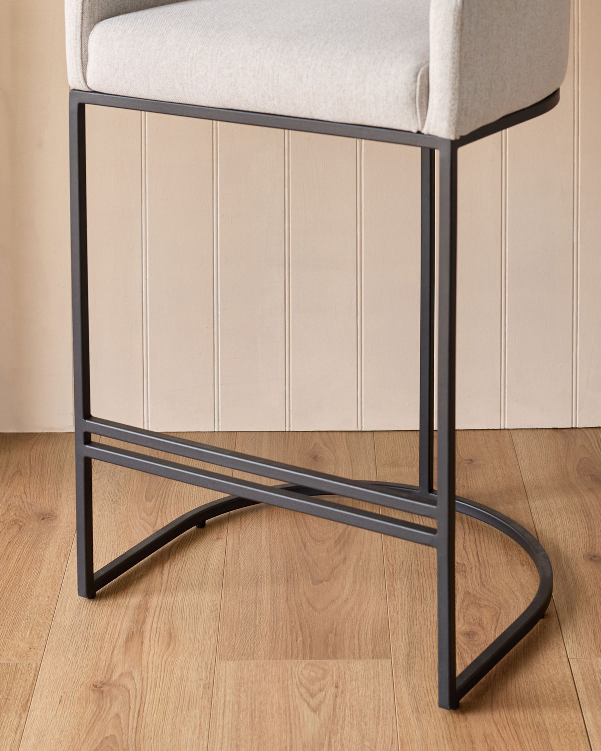 Dakota bar stool with cream fabric seat and black metal frame, shown from side angle on wood flooring.
