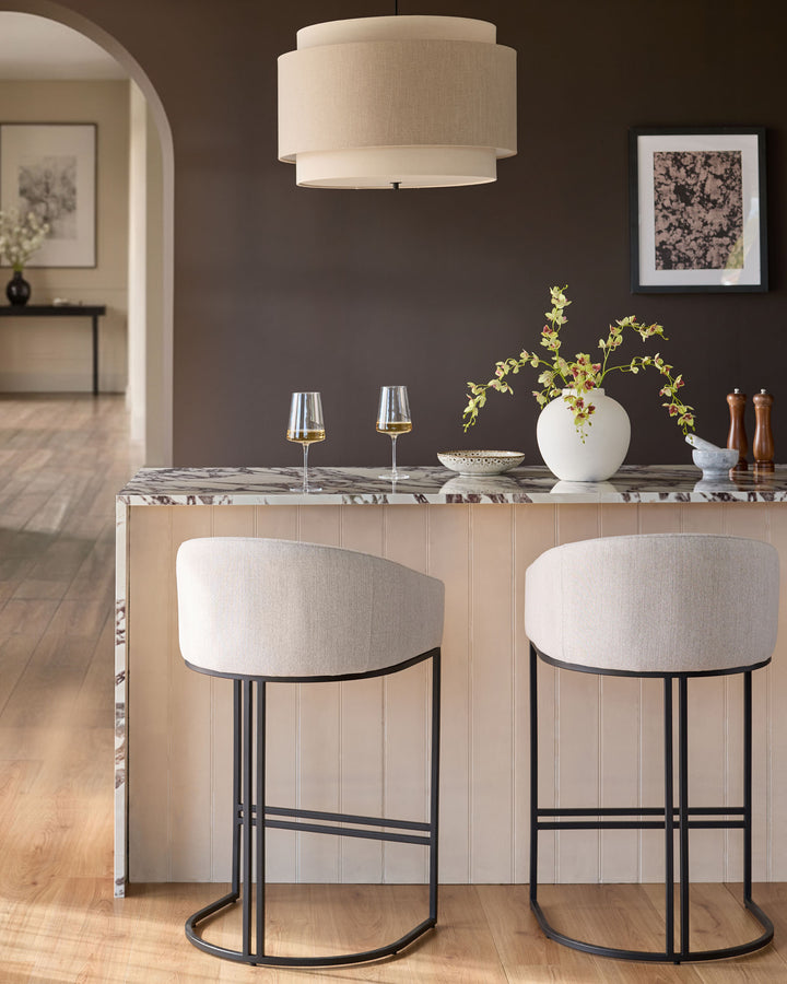 Dakota bar stools in natural fabric with black metal frames, shown as a pair at a marble counter in a modern dining space.