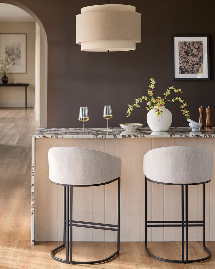 Dakota bar stools in natural fabric with black metal frames, shown as a pair at a marble counter in a modern dining space.