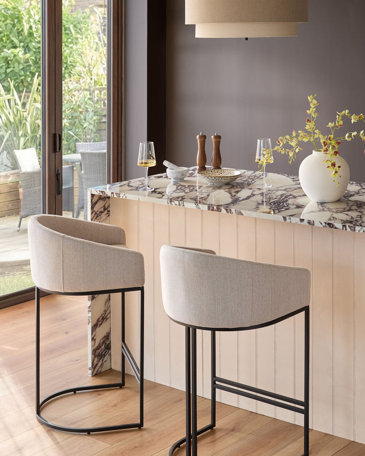 Dakota bar stool in natural fabric with black metal frame, shown at marble-topped kitchen island in modern dining space.