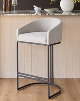 Dakota bar stool in natural fabric with black metal frame, shown against beige paneled wall in modern kitchen setting.