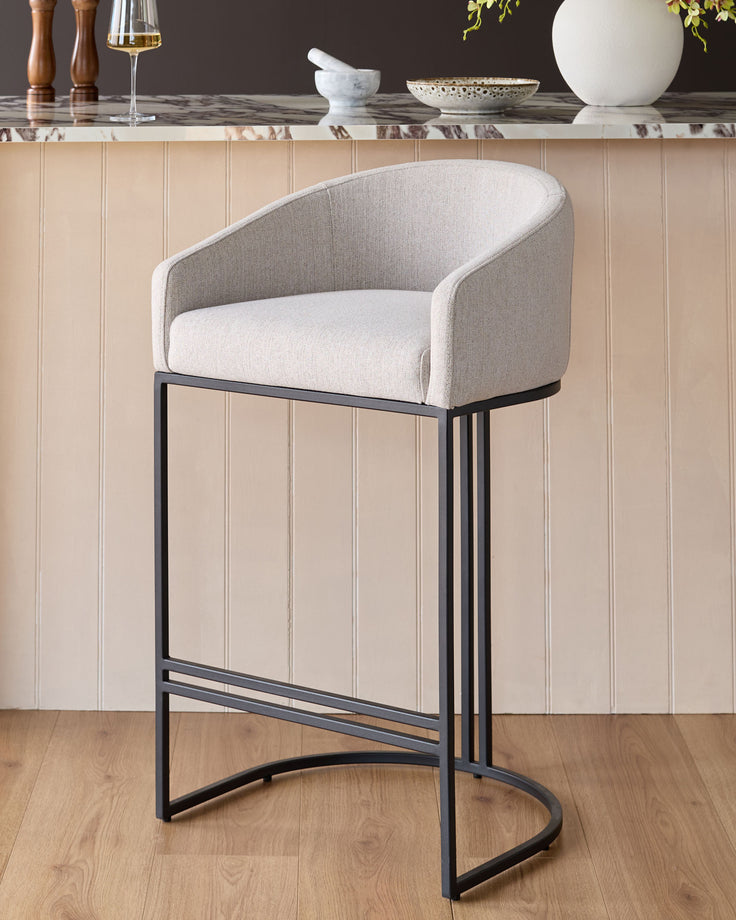 Dakota bar stool in natural fabric with black metal frame, shown against beige paneled wall in modern kitchen setting.