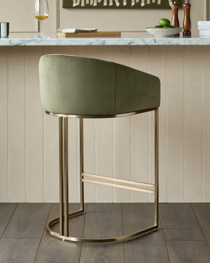 Dakota bar stool in olive green velvet with curved backrest and brushed gold metal frame, shown in kitchen setting.
