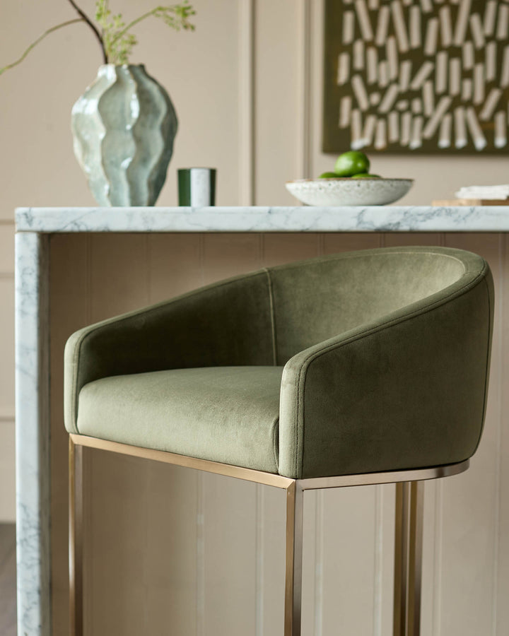 Dakota bar stool in olive green velvet with curved backrest and brushed metal frame, shown at marble countertop.