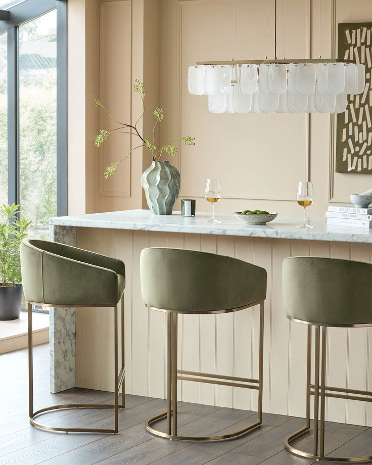 Dakota bar stool in olive green velvet with brushed gold metal frame, shown in set of three at marble countertop.