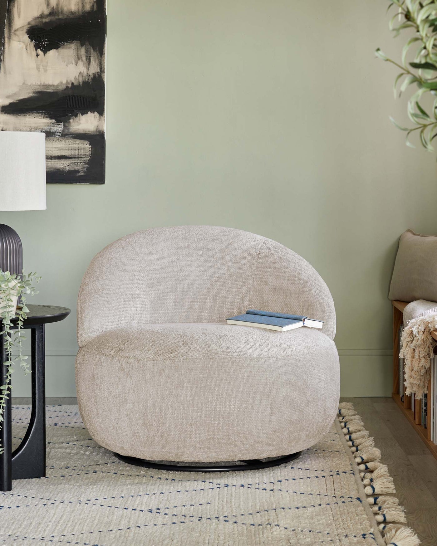 Contemporary beige fabric bouclé accent chair with a rounded, cosy shape, standing on a minimalist black base. Beside it is a small matte black round side table. The room features a woven area rug with tassel details and a cream throw blanket on a wooden bench in the background, creating a warm and inviting space.