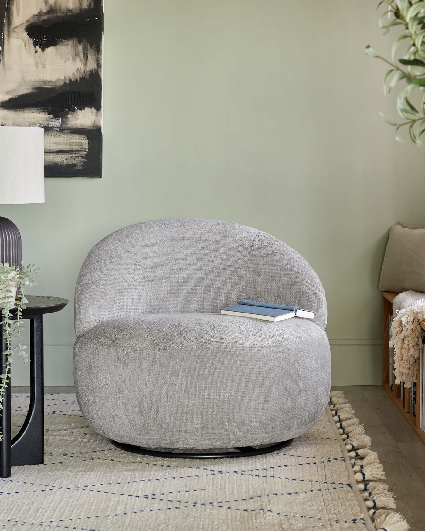 Contemporary grey fabric upholstered curved armchair with a plush seat and backrest, set on a textured off-white area rug with blue stripes and tassel edges, accompanied by a minimalist black round side table with a book on top. The setting includes a neutral beige throw on a wooden bench in the background, a modern abstract wall painting, a white floor lamp, and green houseplants, creating a stylish and cosy ambiance.