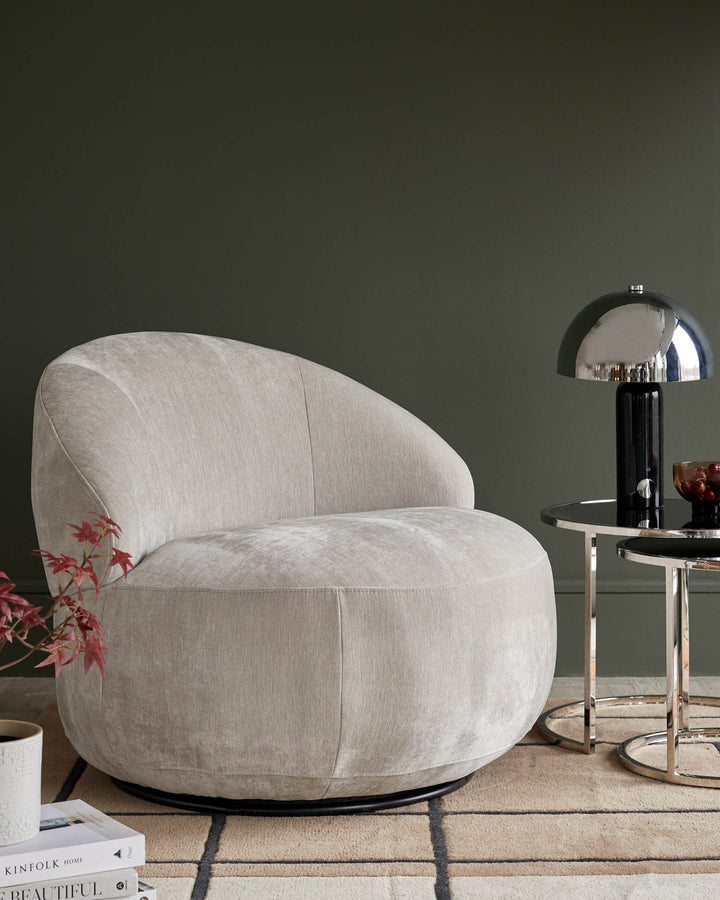 A cosy grey swivel chair next to a chrome and black side table with a lamp and decorative bowl.