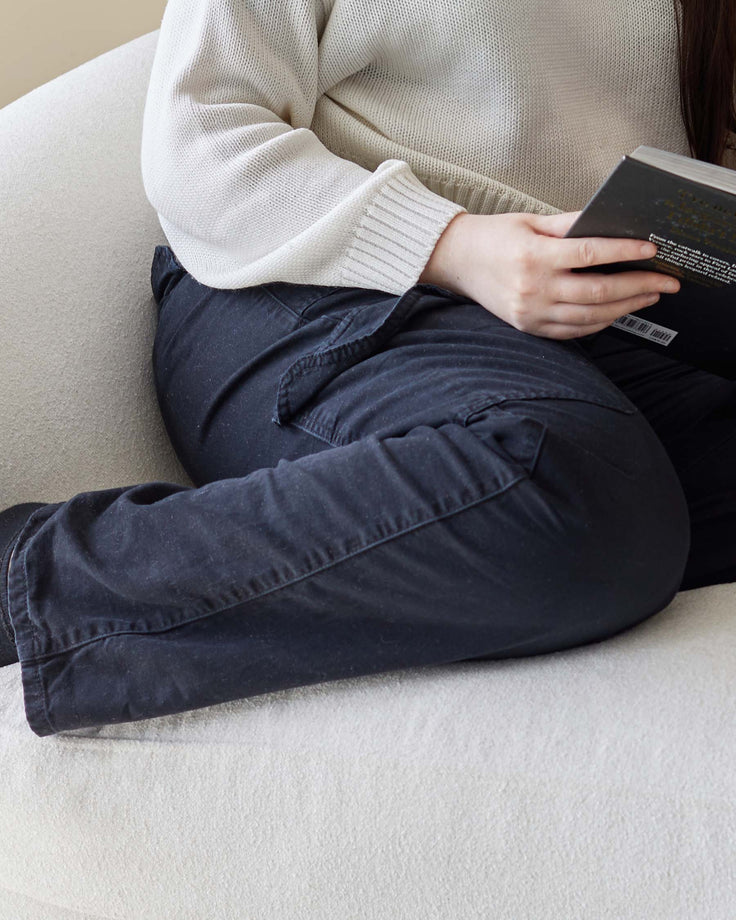 Close-up of a person sat on a white textured sofa, wearing black trousers and a light knit jumper, whilst reading a book.