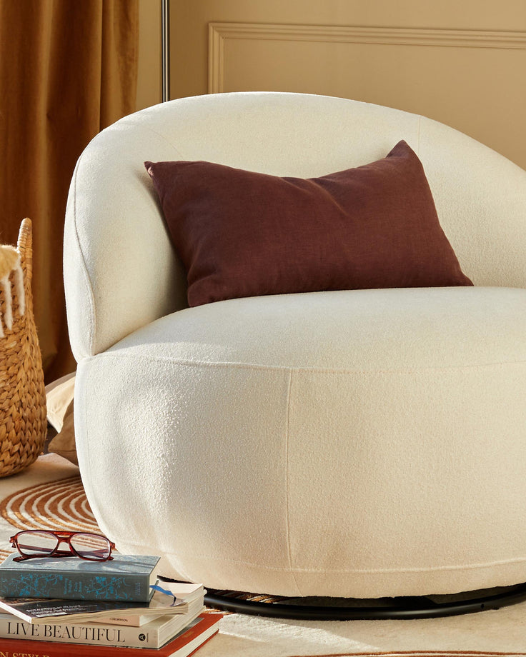 A cream-coloured bouclé swivel chair with a brown cushion, next to a woven basket and stack of books.