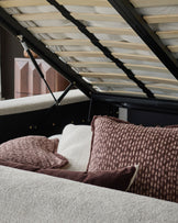 Darla king-size ottoman bed in natural fabric with brown textured pillows, viewed from side angle with open storage mechanism.
