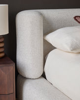 Darla King-Size Ottoman Bed in natural fabric, showing curved cylindrical headboard, white pillows, and wooden nightstand detail.