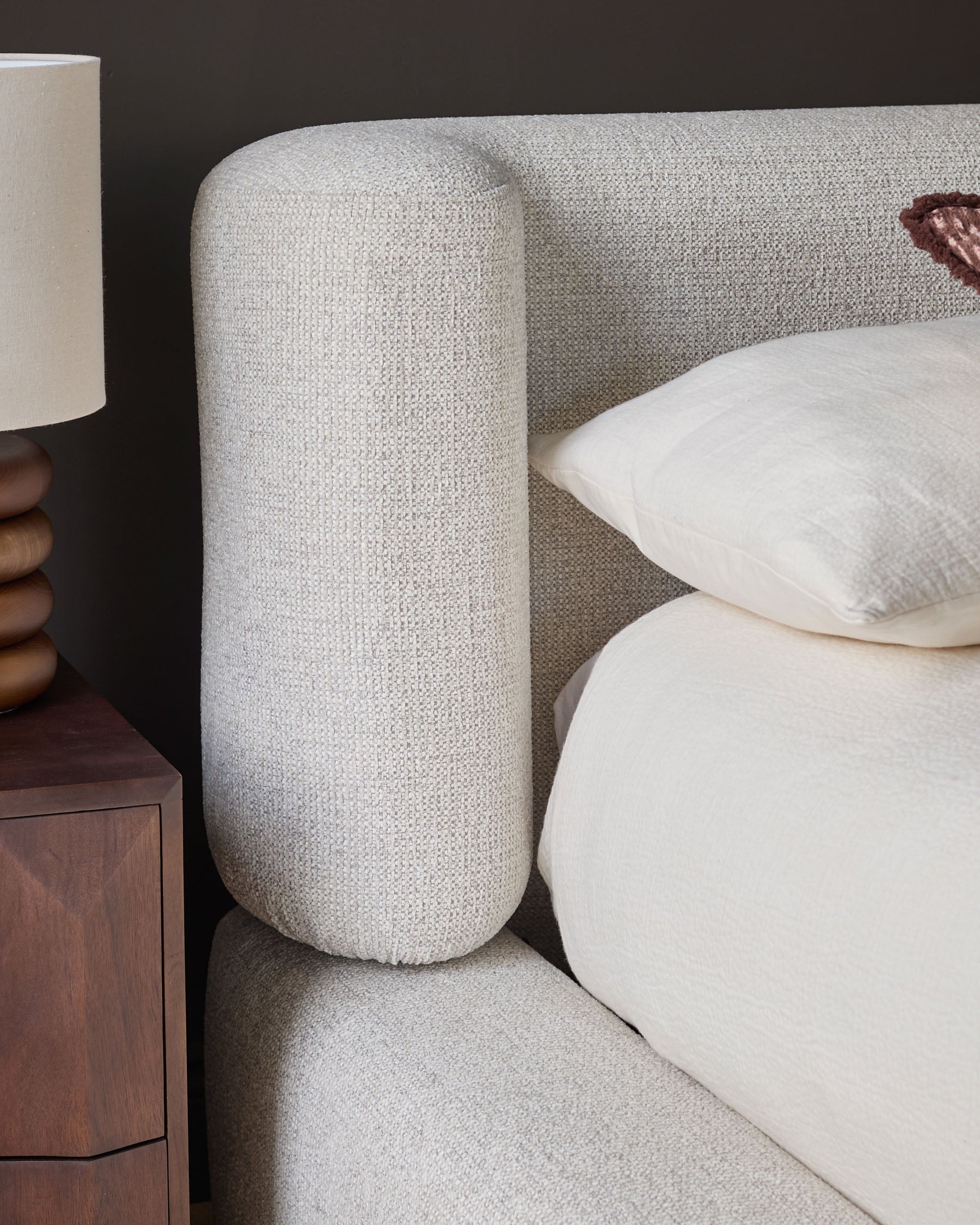 Darla King-Size Ottoman Bed in natural fabric, showing curved cylindrical headboard, white pillows, and wooden nightstand detail.