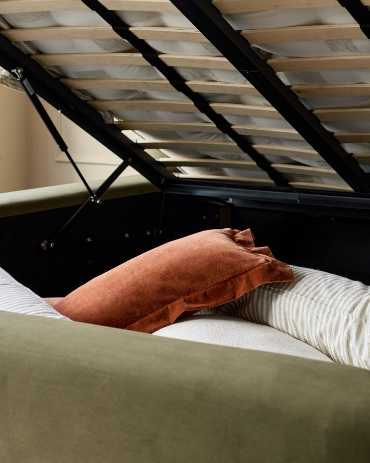 Darla king-size ottoman bed in olive green velvet with open storage base and gas lift mechanism, styled with rust and striped pillows.