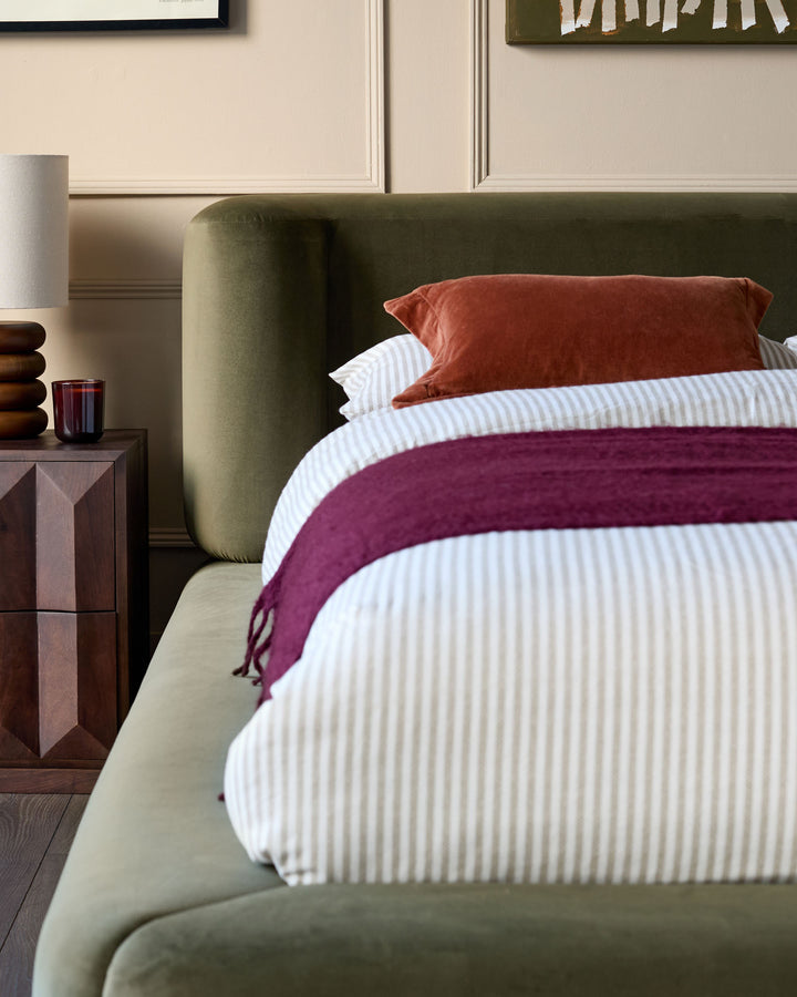 Olive-green velvet ottoman bed with burgundy throw and rust cushions, styled with white striped bedding and wooden nightstand.