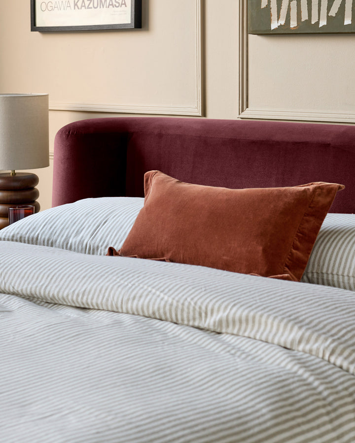 Darla King-Size Ottoman Bed in plum velvet, shown with striped bedding and rust-colored pillow in a styled bedroom setting.