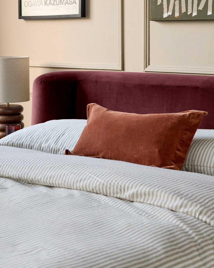Darla King-Size Ottoman Bed in plum velvet, shown with striped bedding and rust-colored pillow in a styled bedroom setting.
