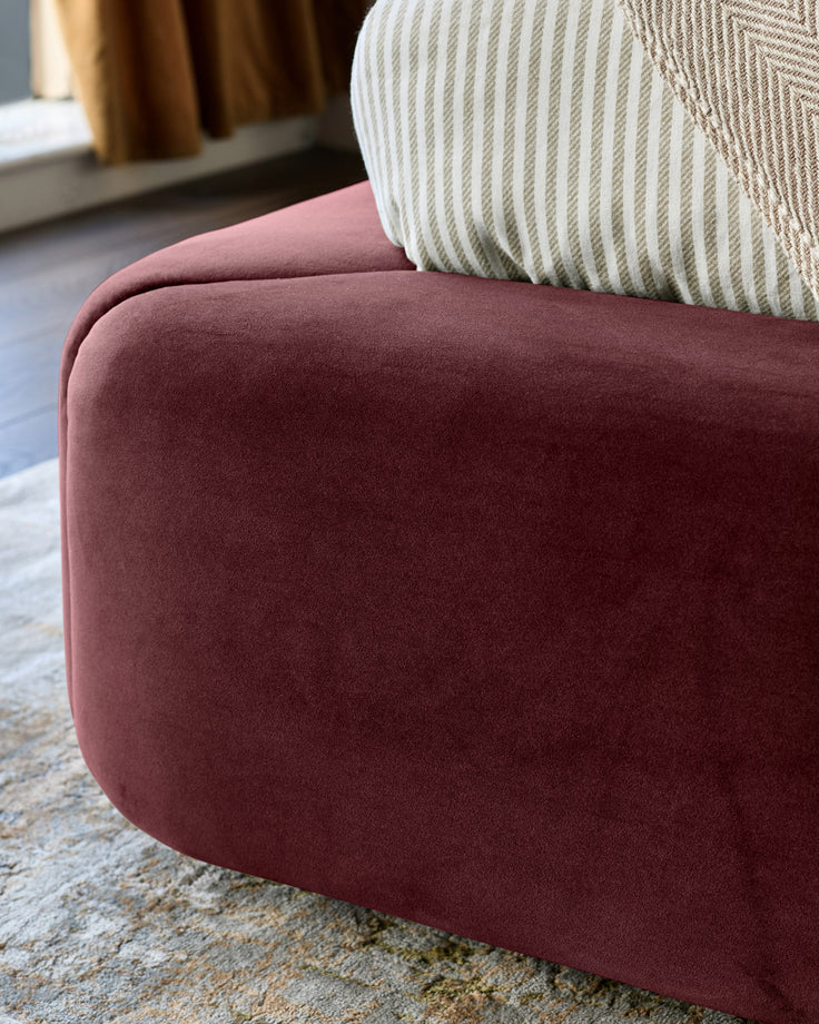 Plum velvet ottoman with rounded edges, striped cream pillow on top, positioned on neutral rug in modern interior.