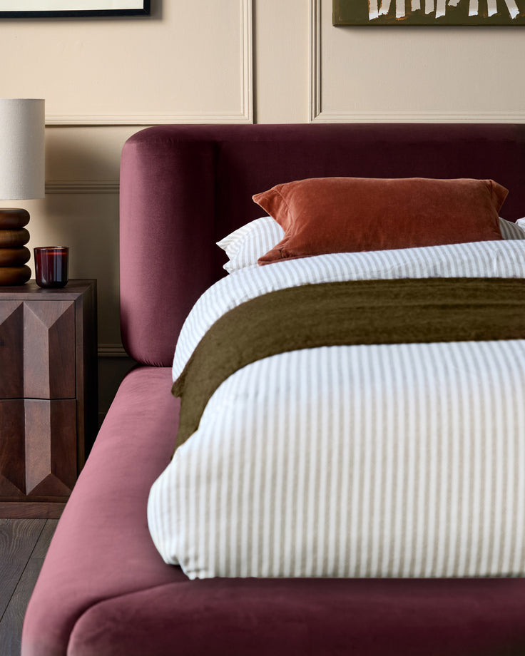 Darla King-Size Ottoman Bed in plum velvet, styled with layered bedding in white and olive green tones, shown from side angle.