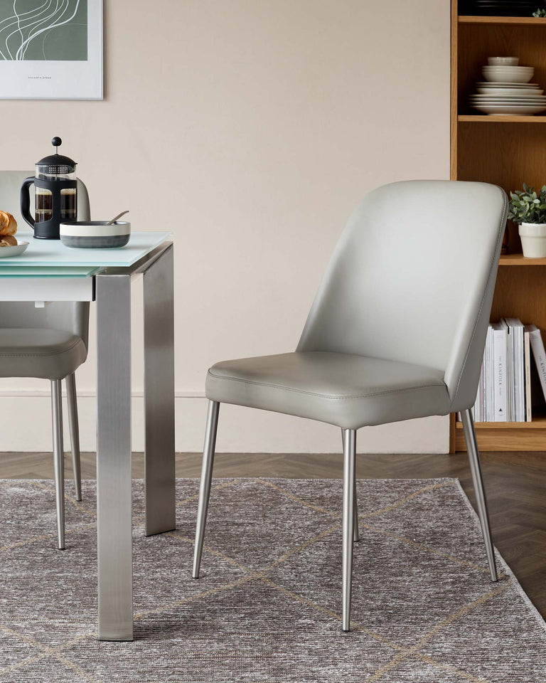 Contemporary light grey upholstered dining chair with a curved back and slim metal legs, paired with a modern glass-top table with a metal frame, set on a grey patterned area rug. A wood shelving unit with dishes and decorative items is partially visible in the background.