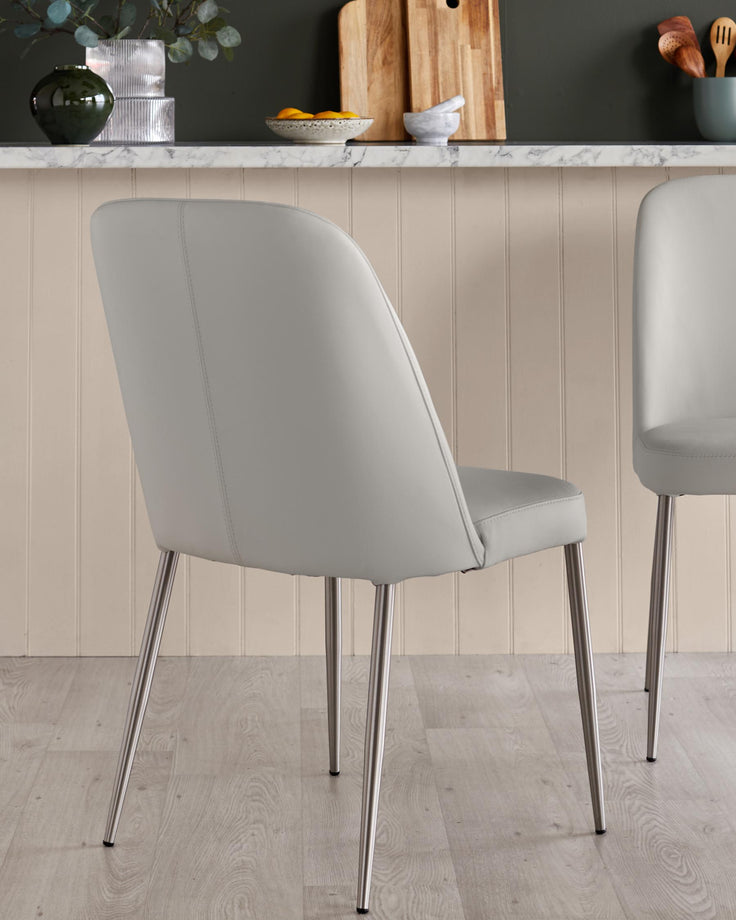 Two modern dining chairs with light grey faux leather upholstery and brushed metal legs, photographed in a kitchen setting.