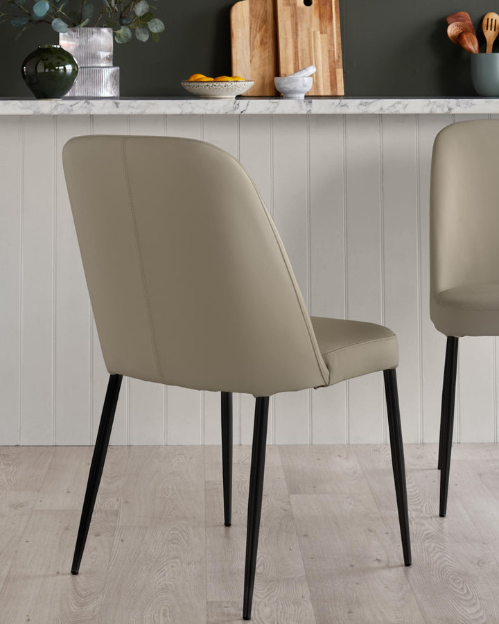 Two taupe dining chairs with black metal legs are arranged against a white panelled wall and marble countertop.