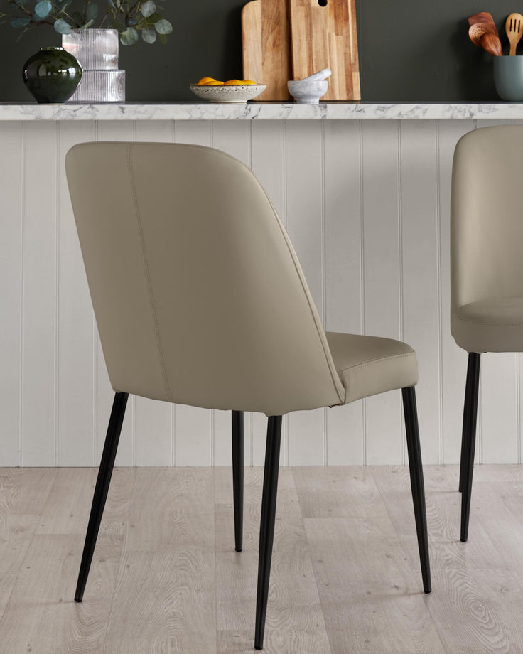Two taupe dining chairs with black metal legs are arranged against a white panelled wall and marble countertop.