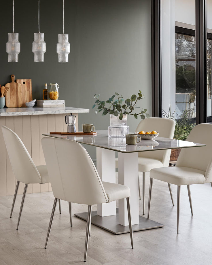 A modern dining set with a glass table, white pedestal, and four cream leather chairs with silver legs.
