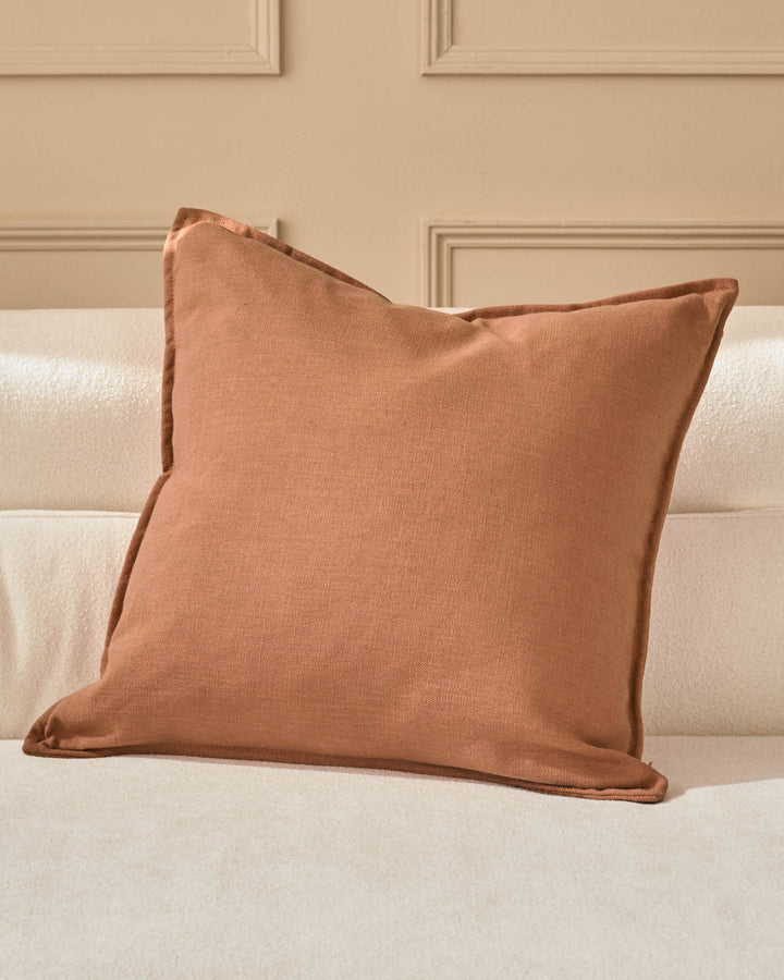 Rust-colored square cushion with piped edges, displayed on a cream upholstered sofa against a beige paneled wall.