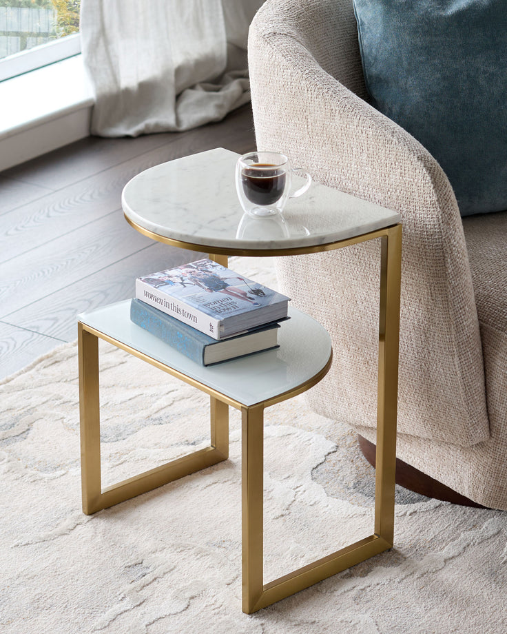Demi side table in white marble with brass frame, shown styled next to a cream armchair with books and coffee cup.