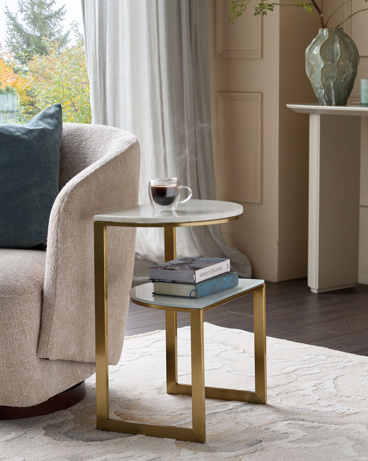 Demi side table in white marble with brushed gold metal frame, shown in a styled living room setting next to a cream armchair.