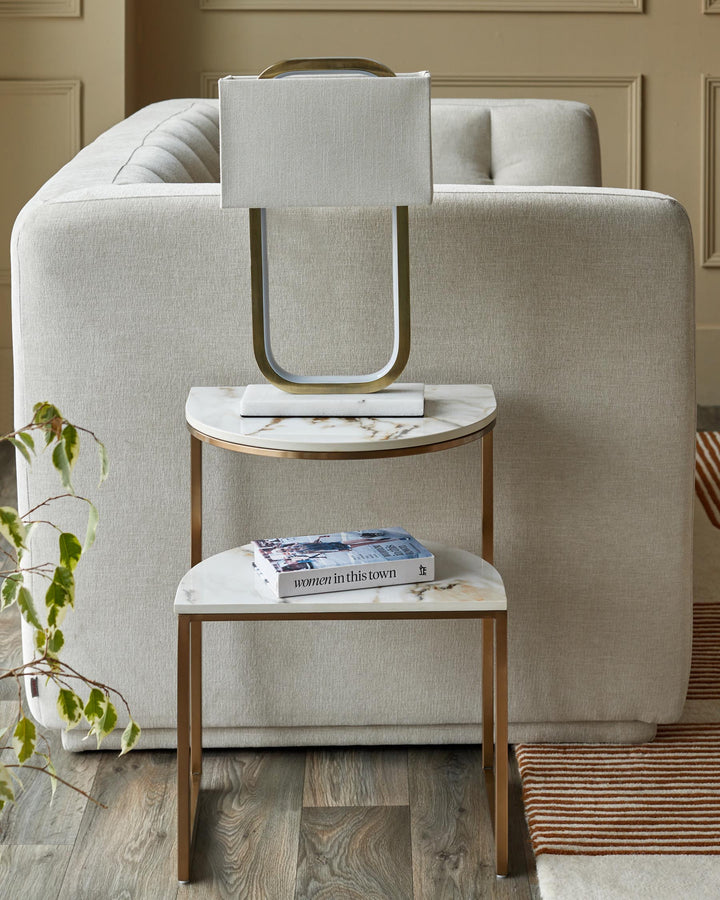 A modern side table with marble tops and gold frame sits beside a cream sofa with a stylish lamp.