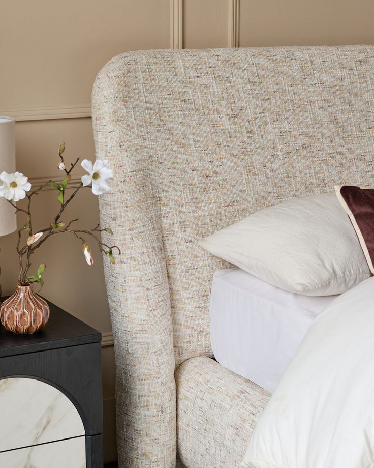 Deven king-size ottoman bed in natural fabric, showing rounded headboard and white bedding, styled with nightstand and flowers.